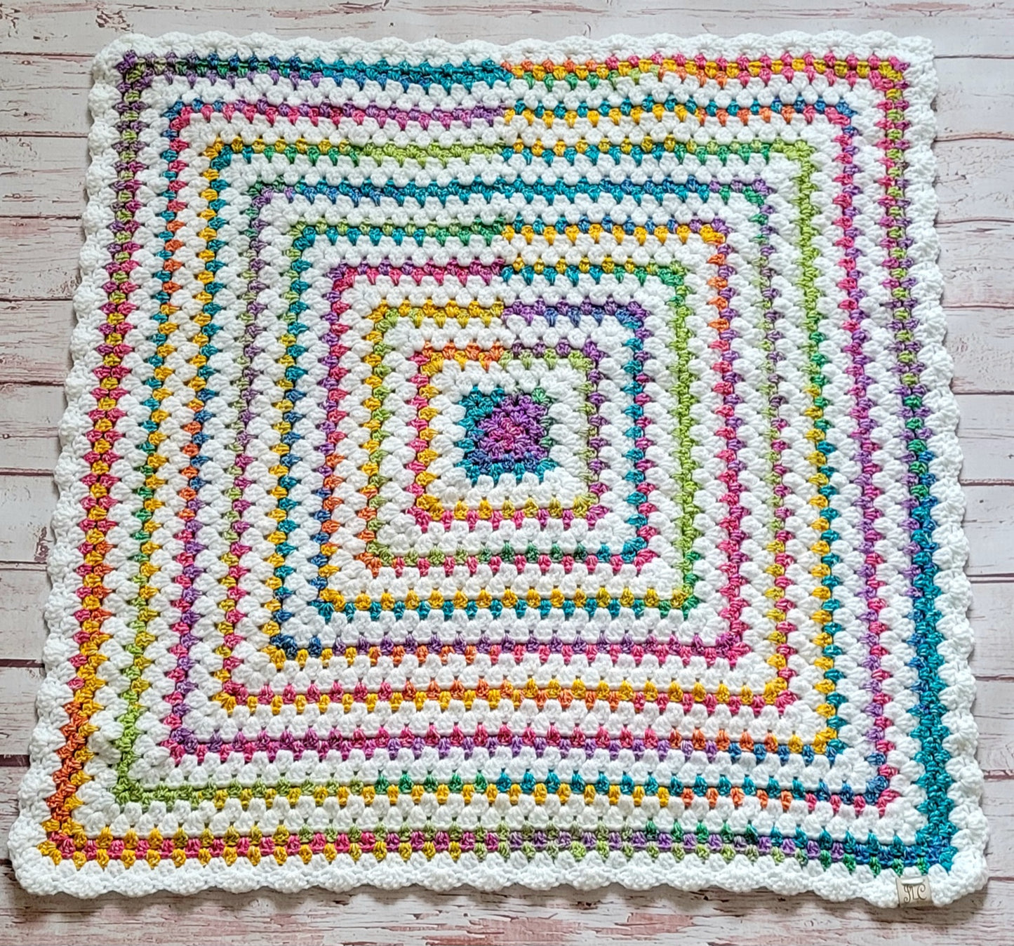 One-of-a-kind 30x30 hand crocheted baby and lap blanket with colorful Rainbow cotton accents, white scalloped border, and Johanna Lesley Creations tag.